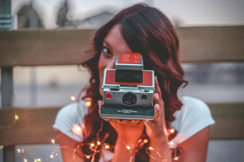 a woman holding a camera in front of her face, a polaroid photo, inspired by Elsa Bleda, pexels contest winner, light red and deep orange mood, bright camera flash enabled, hasselblad film bokeh, holiday