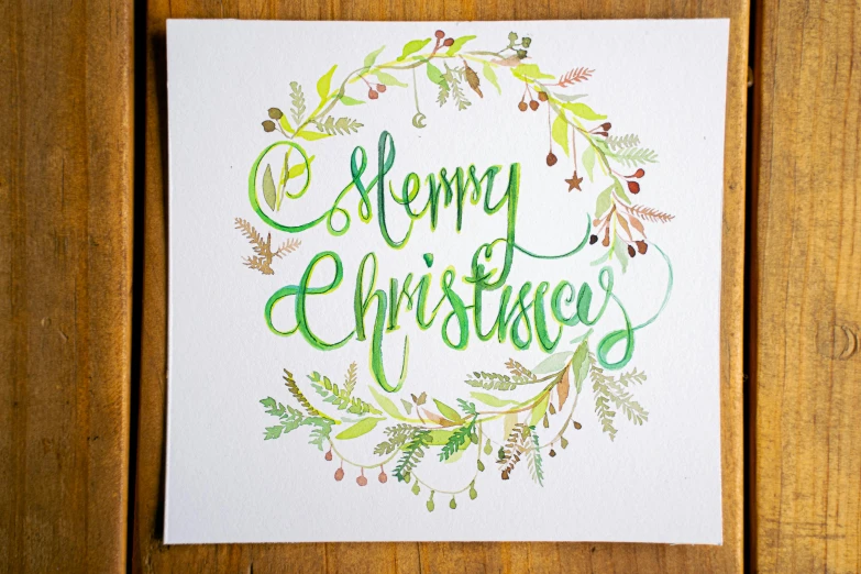 a christmas card sitting on top of a wooden table, a watercolor painting, pixabay, green letters, very very intricate, circular, very crisp