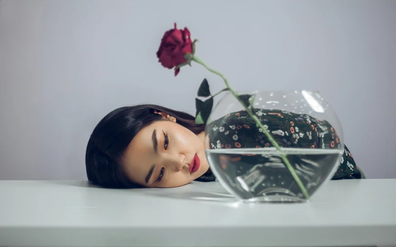 a woman laying down next to a vase with a rose in it, inspired by Fei Danxu, unsplash contest winner, half face in the water, korean girl, charli xcx, sad man