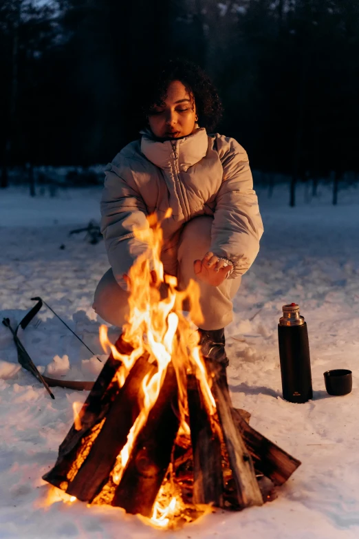 a woman sitting next to a fire in the snow, holding a bottle of arak, imaan hammam, campfire, profile image