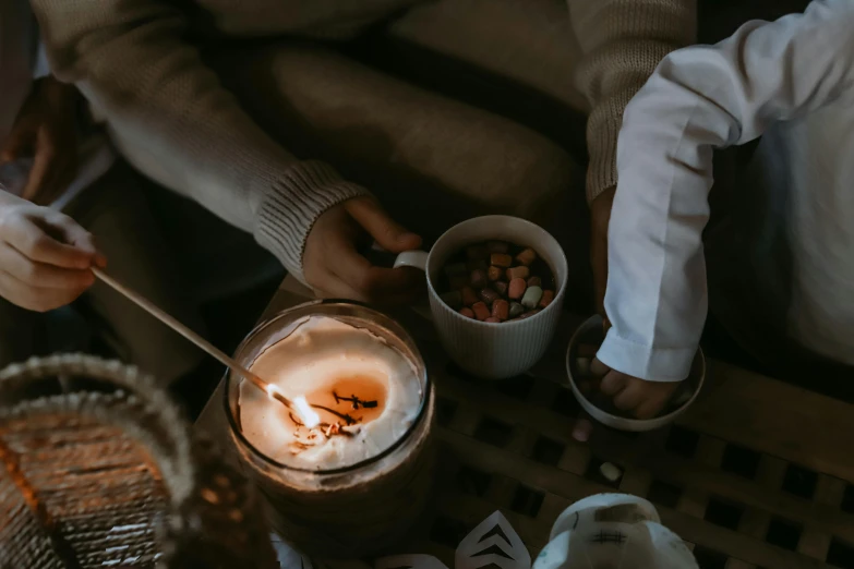 a couple of people sitting at a table with a candle, pexels contest winner, hot cocoa drink, brown and white color scheme, background image, bonfire