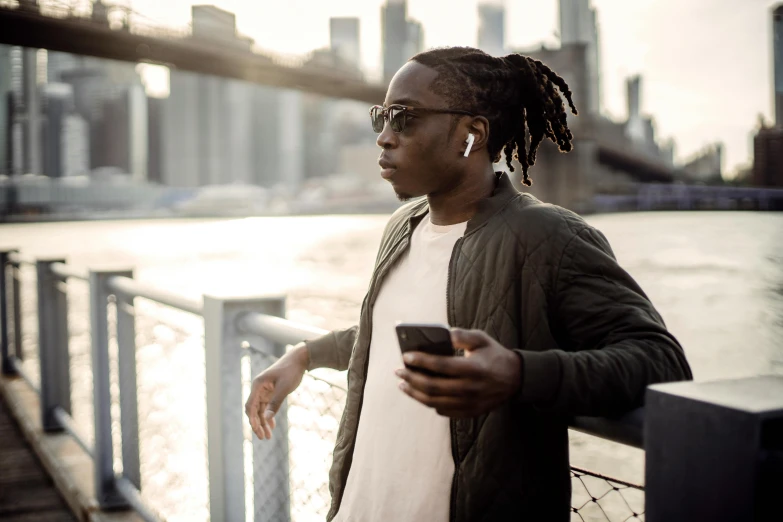 a man standing next to a body of water holding a cell phone, trending on pexels, afrofuturism, dreadlock black hair, implanted sunglasses, humans of new york style, earbuds jewelry