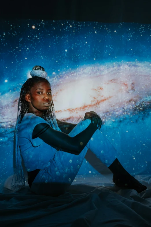 a woman sitting on a bed in front of a picture of a galaxy, an album cover, by Dulah Marie Evans, trending on unsplash, afrofuturism, wears tiny spacesuit, blue dreadlocks, model posing, space high school