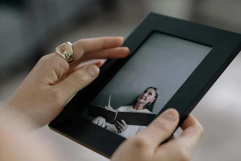 a close up of a person holding a picture in a frame, inspired by Anna Füssli, pexels contest winner, video art, using a magical tablet, reading an evil demonic book, square pictureframes, ultra realistic. cinematic