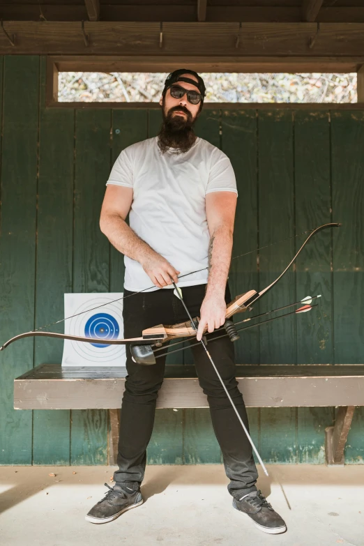 a man sitting on a bench holding a bow and arrow, dustin panzino, beard, standing in an arena, pr shoot