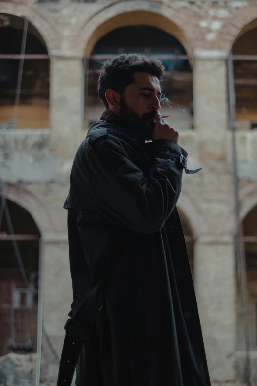 a man standing in front of a building smoking a cigarette, an album cover, by Youssef Howayek, pexels contest winner, renaissance, wearing dark robe, bearded, ( ( theatrical ) ), trench coat with many pockets