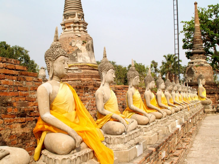 a row of buddha statues sitting on top of a brick wall, with yellow cloths, temples behind her, thai architecture, 🦩🪐🐞👩🏻🦳