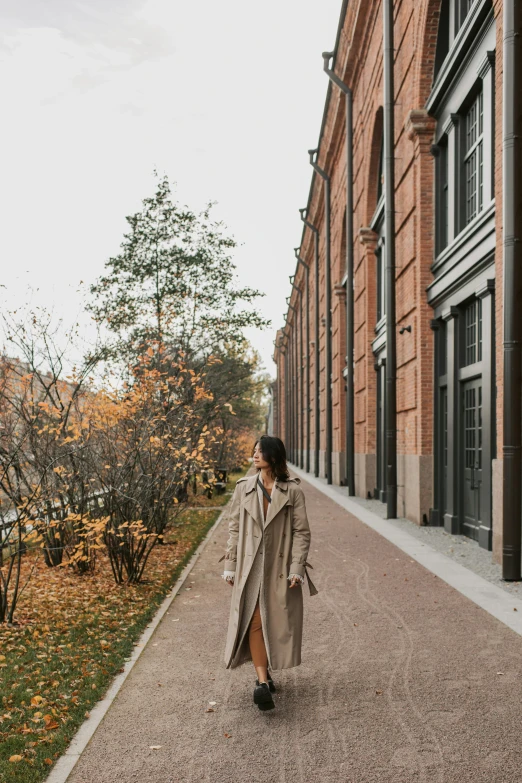 a woman walking down a sidewalk next to a building, inspired by Albert Paris Gütersloh, pexels contest winner, wearing a long coat, brick building, moscow, botanical garden