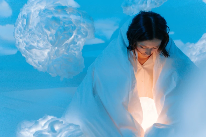 a woman sitting on top of a bed covered in a blanket, interactive art, with ice powers, brightly lit blue room, dressed in white robes, model wears a puffer jacket