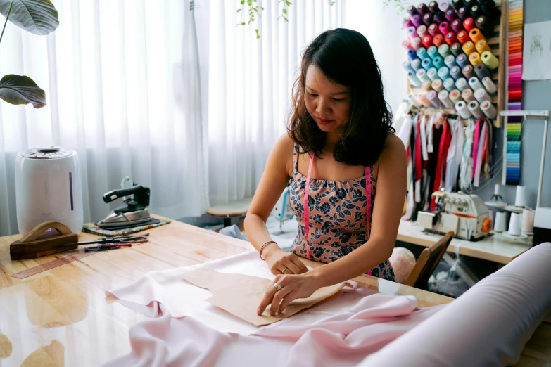 a woman sitting at a table working on a piece of fabric, inspired by Ruth Jên, trending on unsplash, wearing a camisole and shorts, tailored clothing, thumbnail, mai anh tran