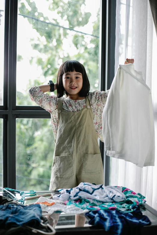 a little girl that is standing in front of a window, laundry hanging, wearing a light shirt, a young asian woman, promo image