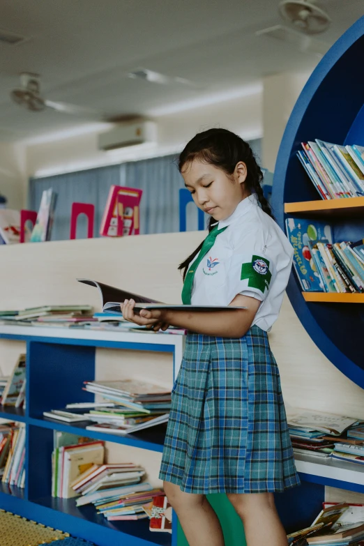 a little girl that is standing in front of a book shelf, wearing a school uniform, reading, thailand, in pif