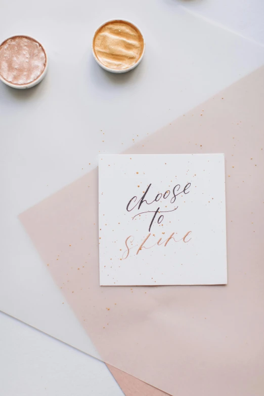 a card sitting on top of a table next to a cup of coffee, by Lucette Barker, trending on unsplash, rose gold, sparklers, monochromatic colour palette, made of silk paper