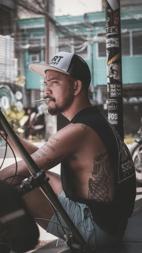 a man sitting on the ground smoking a cigarette, a tattoo, inspired by Eddie Mendoza, pexels contest winner, sleeveless, sitting on a motorcycle, profile image, caracter with brown hat