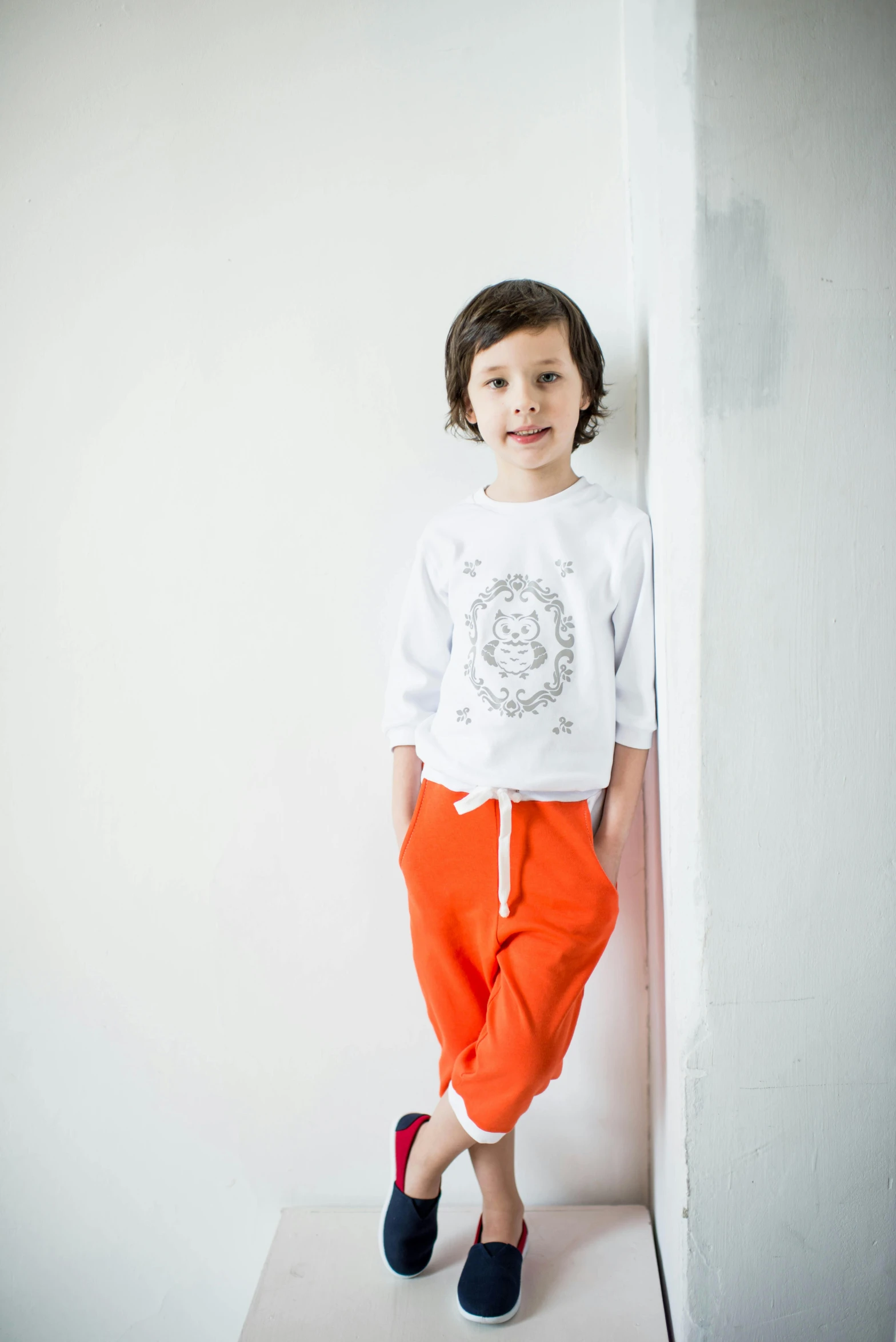 a little boy that is standing up against a wall, inspired by Louis Le Nain, wearing an orange t shirt, in a white boho style studio, wearing a tracksuit, bright white light