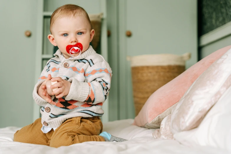 a baby sitting on a bed with a pacifier in his mouth, a portrait, by Julia Pishtar, pexels, crimson themed, bowater charlie and brom gerald, casually dressed, confident looking