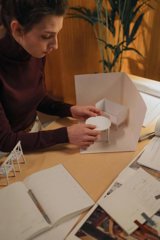 a woman sitting at a table working on a project, by Mathias Kollros, paper craft, architectural presentation, promo image, assembly instructions