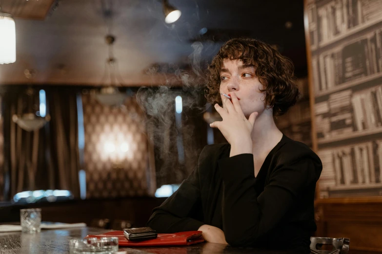 a man sitting at a table with a cigarette in his mouth, by Julia Pishtar, pexels contest winner, attractive girl tomboy, finn wolfhard, sitting at the bar, an elegant