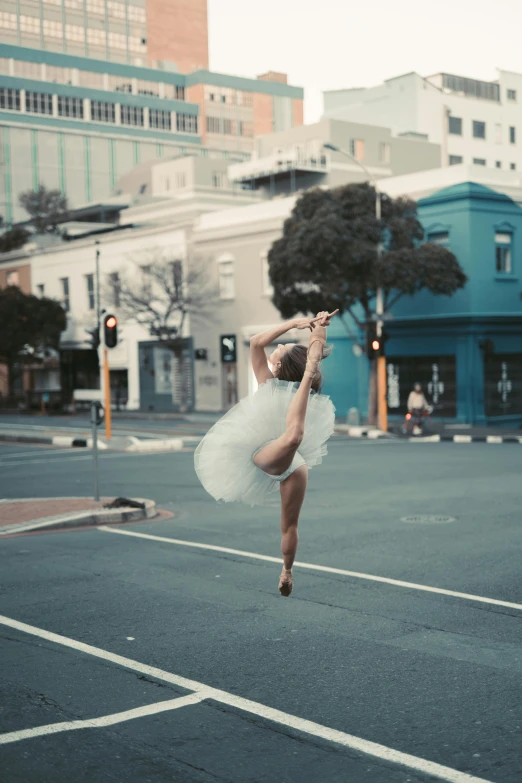 a woman in a tutu jumping in the air, by Lee Loughridge, unsplash contest winner, arabesque, photorealistic streetscape, square, blueish tones, intersection