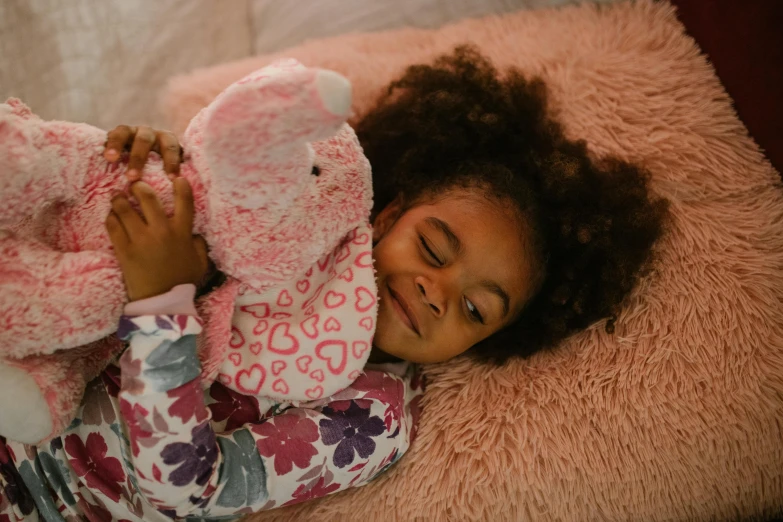a little girl that is laying down with a stuffed animal, inspired by Sophie Anderson, pexels, african american girl, pink, bedhead, paisley