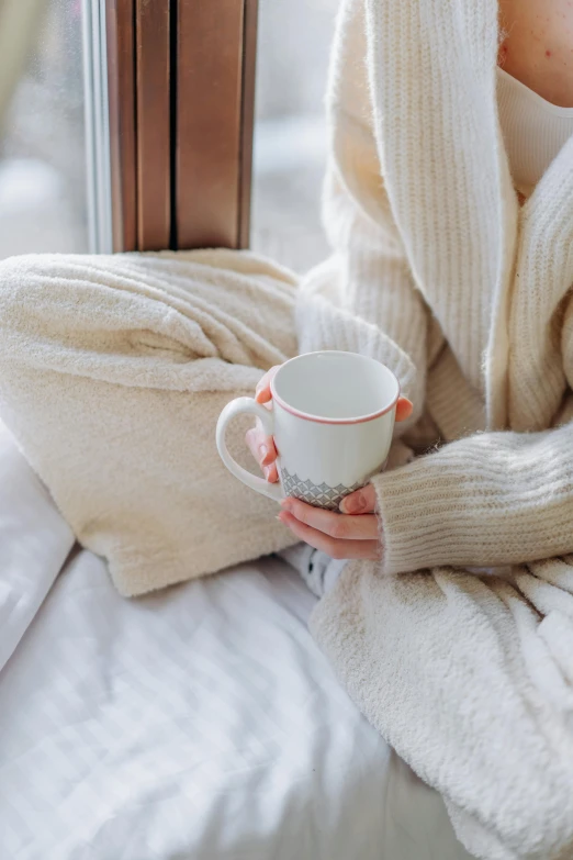 a woman sitting on a bed holding a cup, by Elaine Hamilton, trending on pexels, cold weather, soft outline, folded arms, vanilla