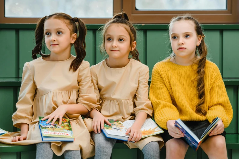 three little girls sitting on a bench with books, inspired by Elsa Beskow, pexels contest winner, danube school, yellow and olive color scheme, sitting in a waiting room, serious focussed look, russian academic