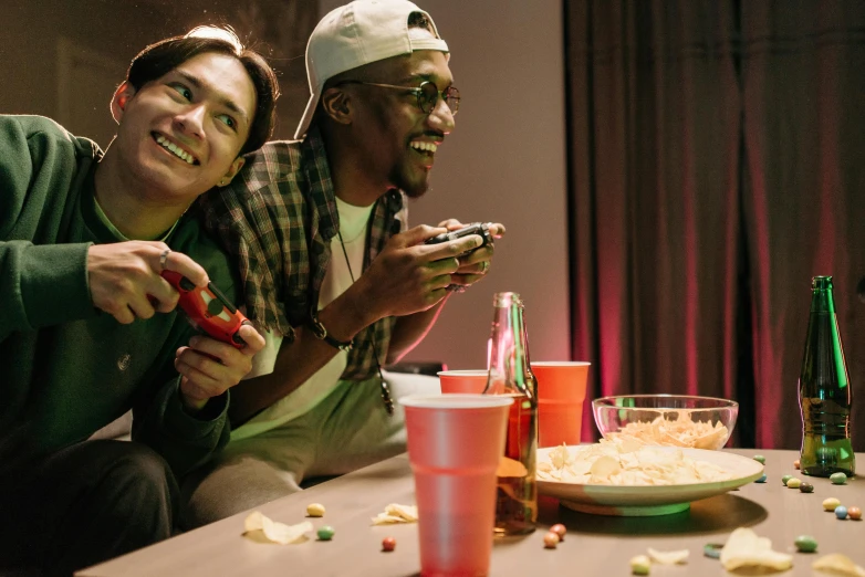 a couple of men sitting next to each other on a table, pexels, nintendo game, eating chips and watching tv, having a cool party, mix of ethnicities and genders