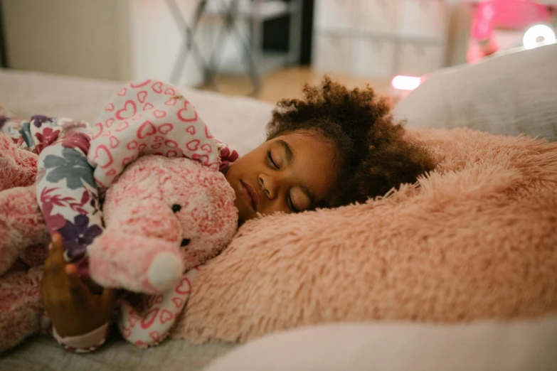 a little girl laying on top of a bed next to a stuffed animal, pexels contest winner, african american girl, brown and pink color scheme, evening time, bedhead