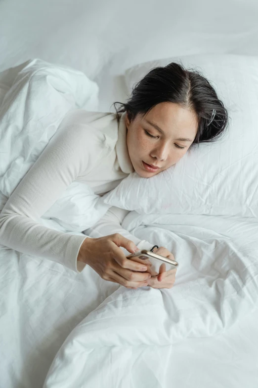 a woman laying in bed with a cell phone, inspired by Ruth Jên, trending on pexels, happening, sleek white, asian woman, high angle, isolation