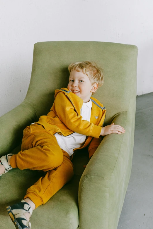 a little boy sitting on top of a green chair, black and yellow tracksuit, on a couch, wearing a yellow hoodie, ocher details