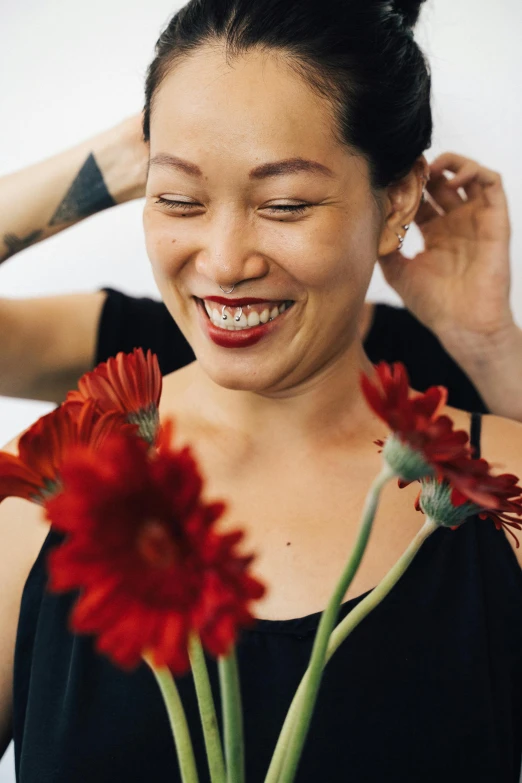 a woman holding a flower in front of her face, an album cover, inspired by Ruth Jên, trending on unsplash, smiling at each other, shaved hair, red flowers, head bent back in laughter