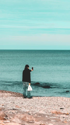 a man standing on top of a beach next to the ocean, smartphone photo, on a canva, holding a stuff, human staring blankly ahead