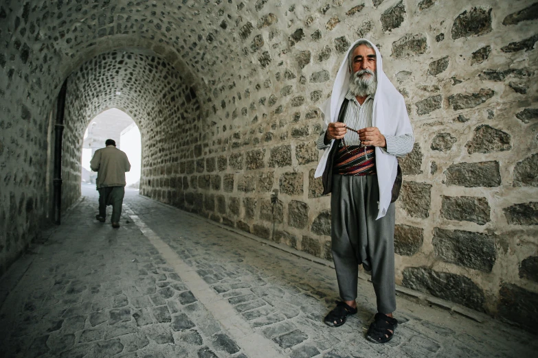 a man standing in front of a stone wall, by Ibrahim Kodra, unsplash contest winner, les nabis, high walled tunnel, two old people, dressed thobe, citadel of erbil