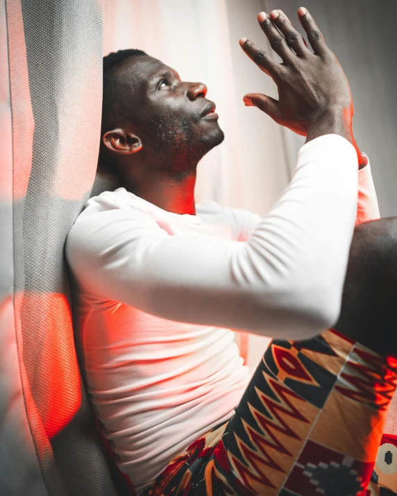 a man in a white shirt leaning against a wall, an album cover, inspired by Paul Georges, pexels contest winner, happening, red light from some windows, sitting on top of a rainbow, ( ( dark skin ) ), grey skinned