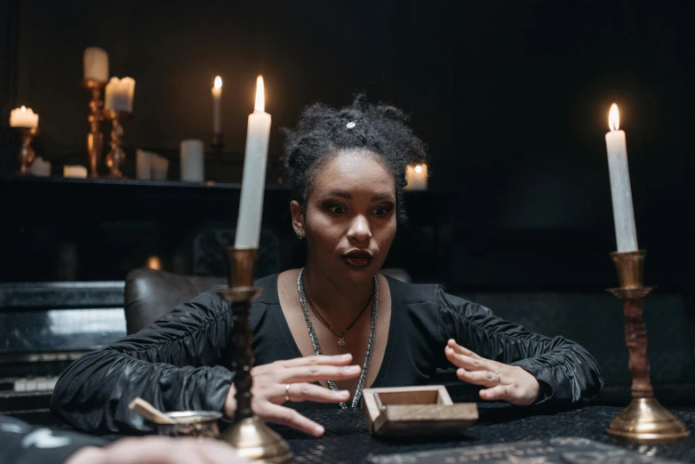 a woman sitting at a table with candles in front of her, inspired by Carrie Mae Weems, trending on pexels, renaissance, casting an evil spell, black young woman, person in foreground, a magician's chamber