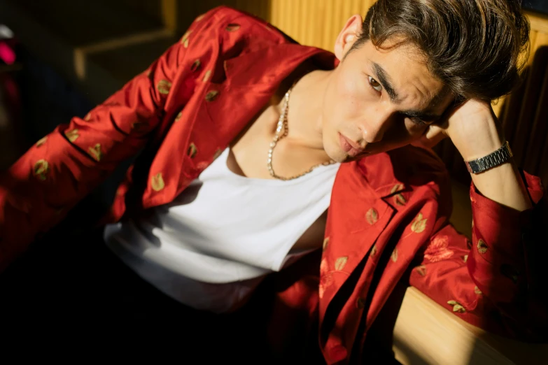 a man in a red jacket sitting on a bench, an album cover, trending on pexels, beautiful androgynous prince, diego fernandez, gold clothes, model pose