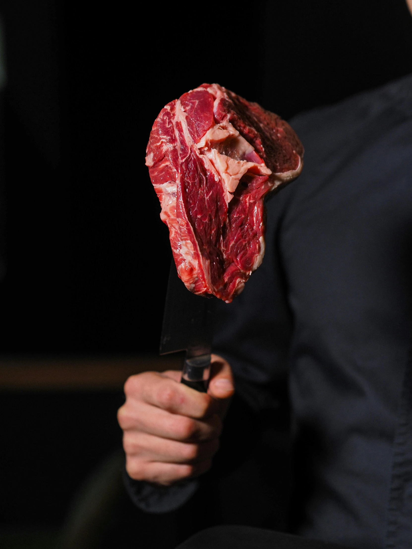 a man holding a knife with a piece of meat on it, hanging, half man half angus bull, against a deep black background, looking to the side off camera