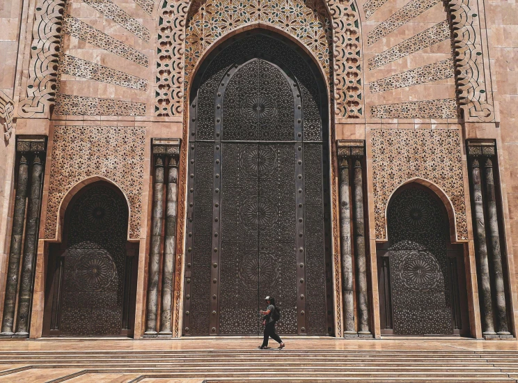 a man walking in front of a large building, inspired by Alberto Morrocco, pexels contest winner, arabesque, huge gate, black and terracotta, islamic, glossy surface