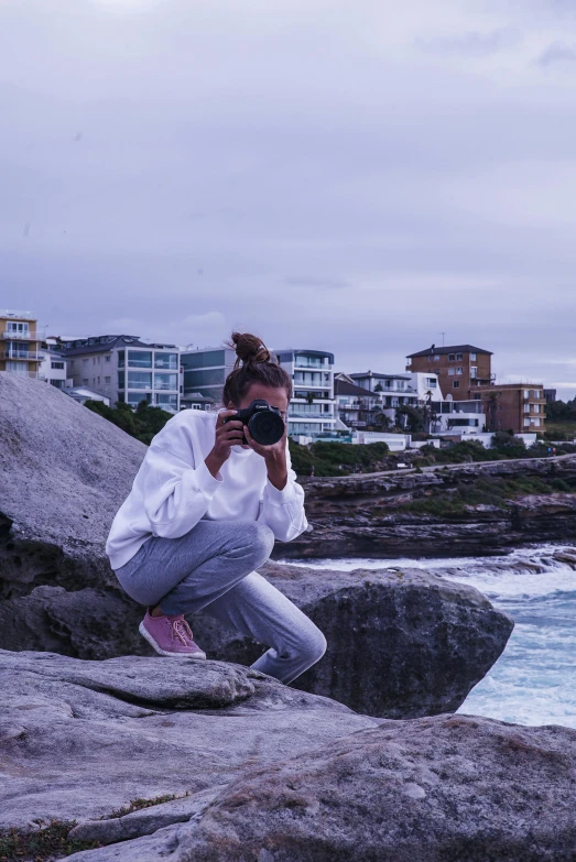 a woman sitting on a rock taking a picture of the ocean, inspired by Sydney Carline, unsplash, photorealism, wearing a purple sweatsuit, bondi beach in the background, camera flash, doing a sassy pose