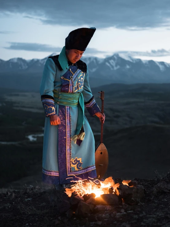 a man standing on top of a hill next to a fire, by Alexander Runciman, unsplash contest winner, cloisonnism, embroidered robes, russian costume, cooking it up, with mountains as background