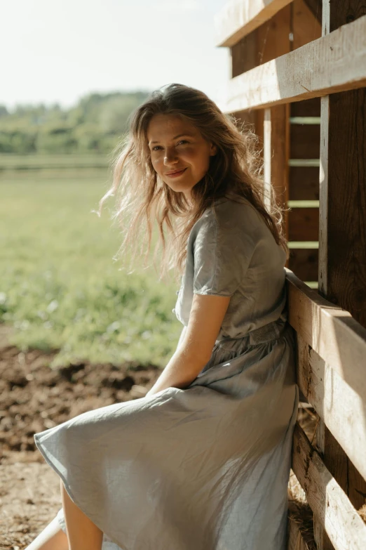 a woman sitting on the side of a wooden fence, profile image, barn in the background, wearing silver dress, greta thunberg smiling