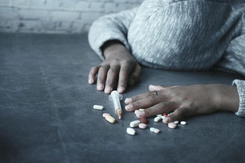 a woman sitting at a table with pills and a cigarette, by Arabella Rankin, trending on pexels, laying down with wrists together, on grey background, head down, multi - coloured