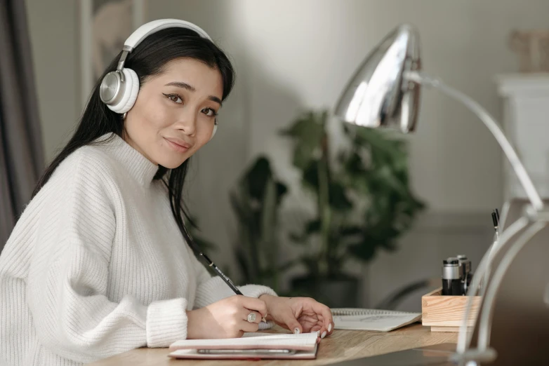 a woman sitting at a desk with headphones on, inspired by Li Di, trending on pexels, academic art, asian female, casually dressed, white hue, lovingly looking at camera