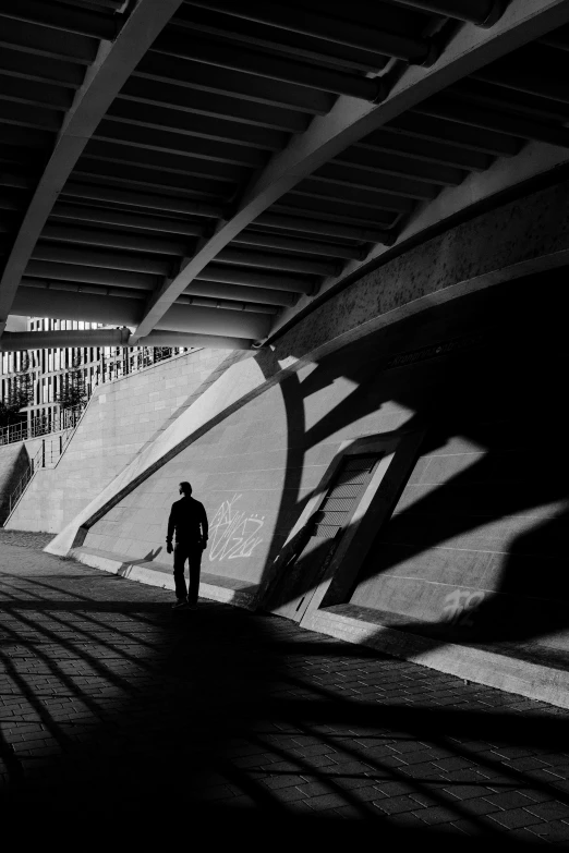 a black and white photo of a man walking under a bridge, deep shadows and colors, london south bank, contre - jour, solitude