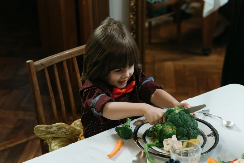 a little girl sitting at a table with a plate of broccoli, by Emma Andijewska, pexels contest winner, finn wolfhard, cutting a salad, very handsome, hotei is on the table