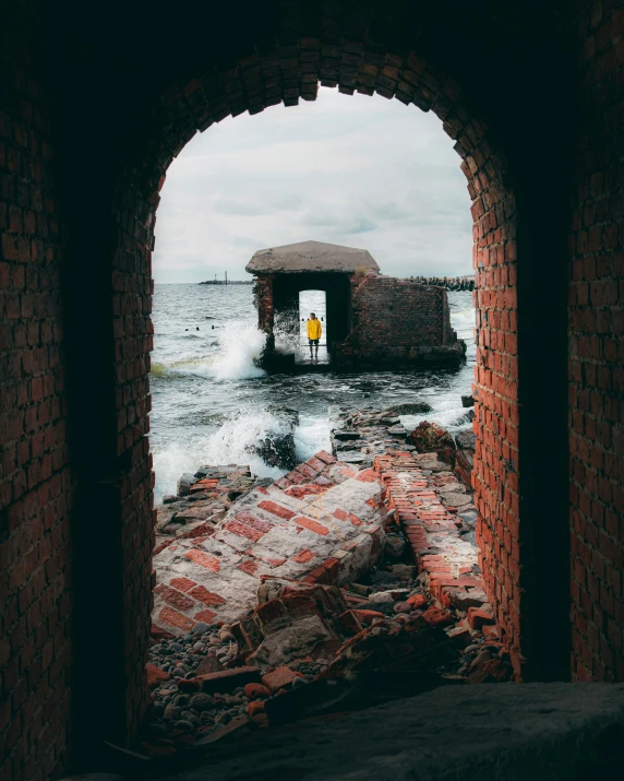 a man standing on top of a rock next to the ocean, a picture, pexels contest winner, visual art, arched doorway, brick building, damaged photo, inside on a rainy day