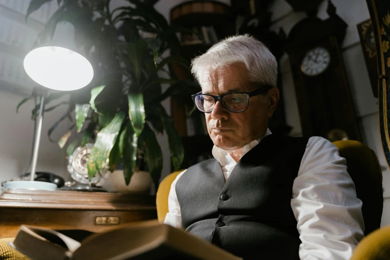 a man sitting in a chair reading a book, a character portrait, inspired by John Watson Gordon, pexels, photorealism, a silver haired mad, reading glasses, low - lighting, cinematic movie still