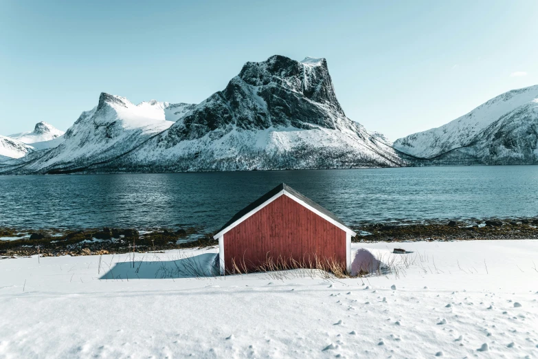 a red building sitting in the snow next to a body of water, by Brian Snøddy, pexels contest winner, tall mountains in the horizon, whitewashed housed, cottage, a photo of the ocean