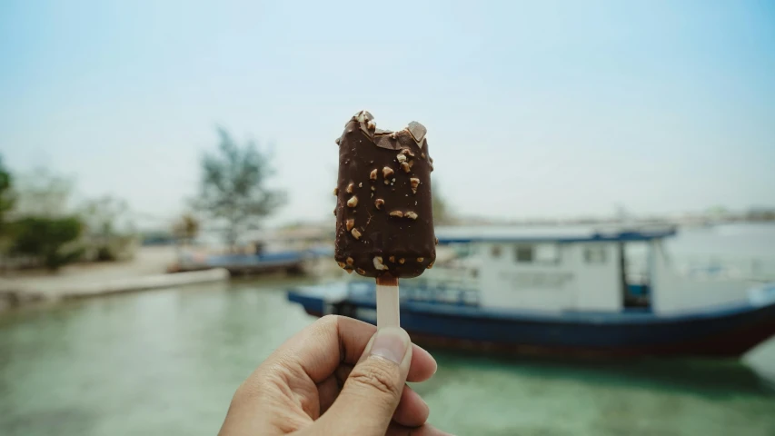 a close up of a person holding a popsicle near a body of water, fully chocolate, singapore, boat, high quality image