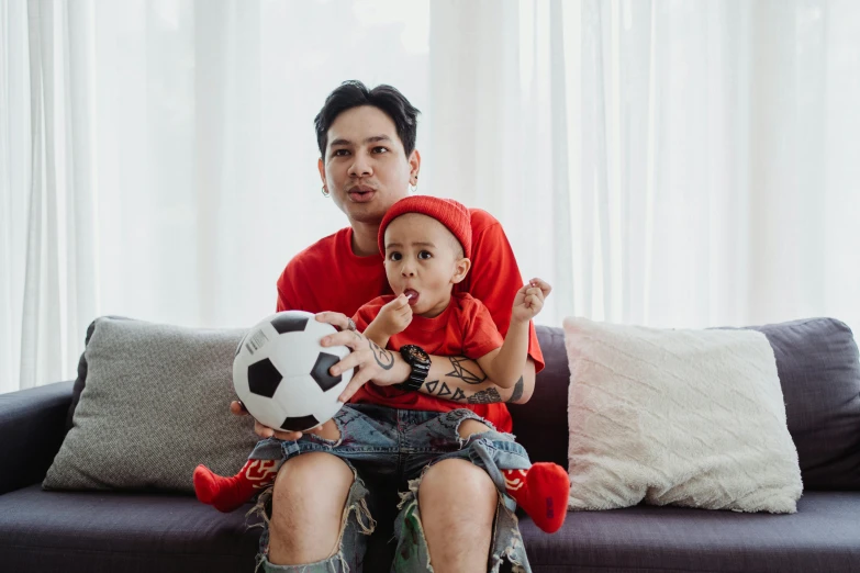 a man sitting on a couch with a baby holding a soccer ball, a cartoon, by Julia Pishtar, pexels contest winner, wearing red attire, hoang long ly, fatherly, lgbtq